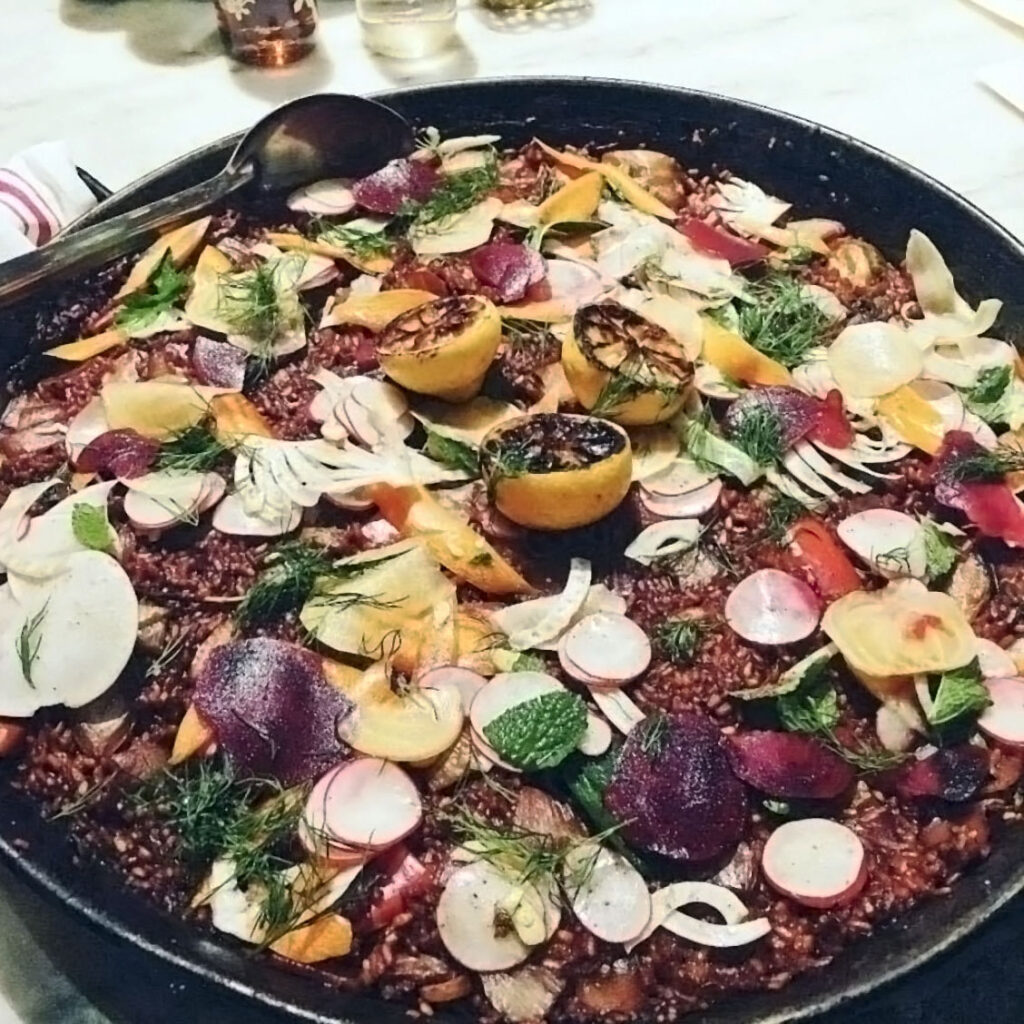 Colorful vegetables and a spoon in a pan of Roasted Beet Paella on a table.