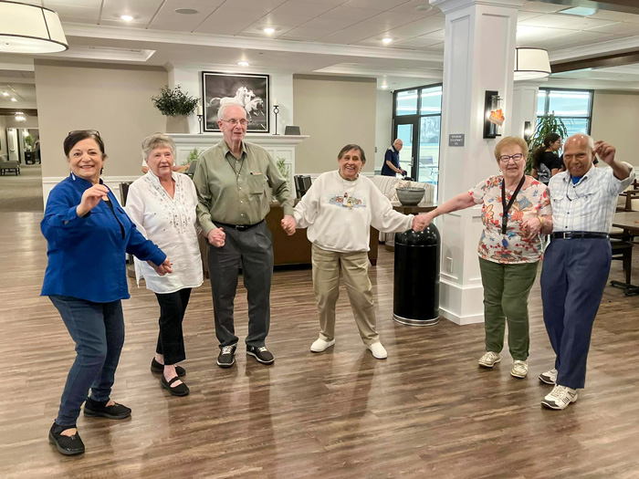 A joyful group of six seniors holding hands and dancing together indoors, capturing the spirit of community, friendship, and active living in a vibrant senior living environment.