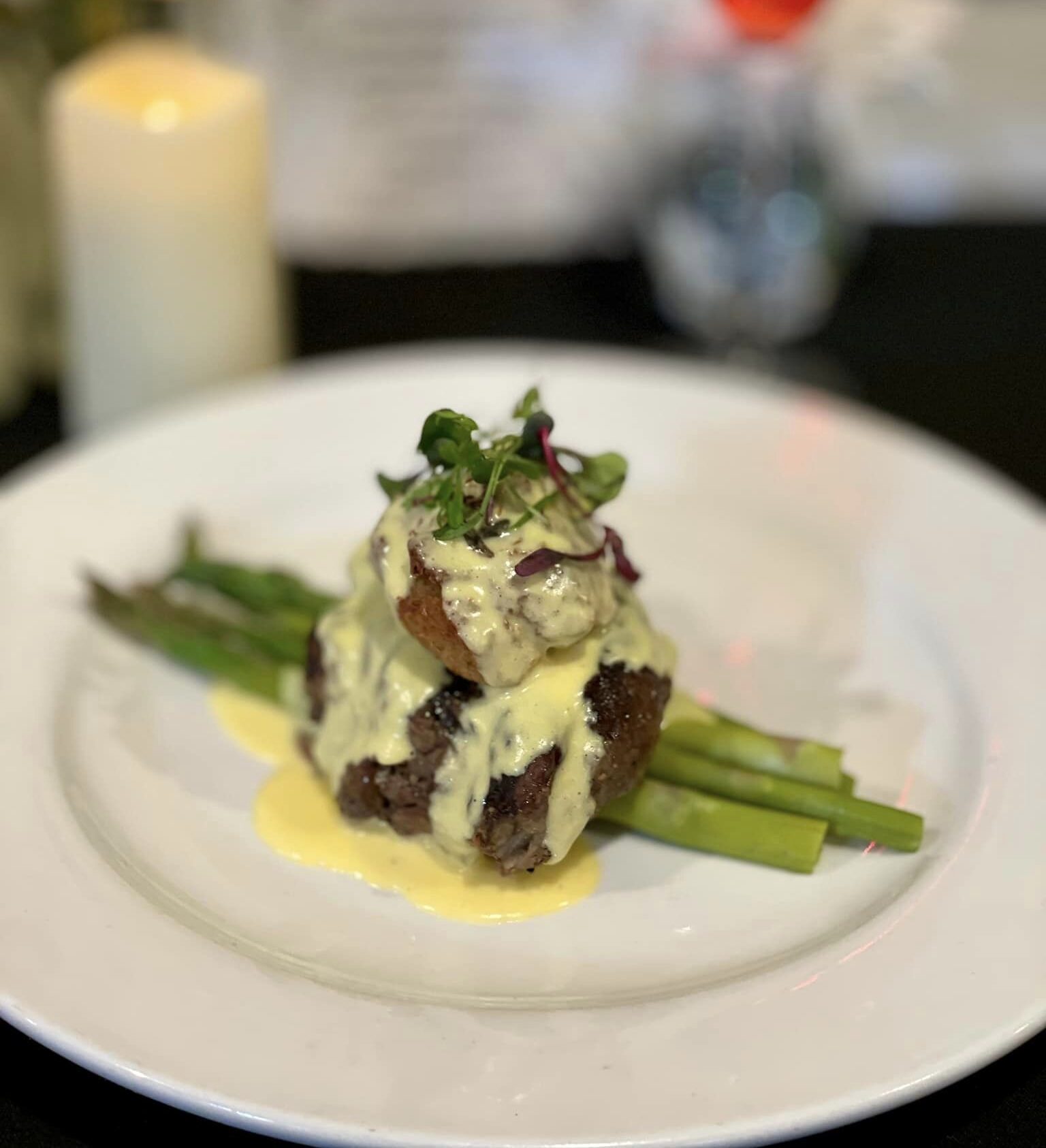 Plated gourmet steak with asparagus, topped with creamy hollandaise sauce and garnished with microgreens, set against a blurred background of a candle, flowers, and drink.