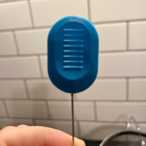 Close-up of a handheld blue digital food thermometer with a ridged grip, held in front of a white subway tile kitchen backsplash.