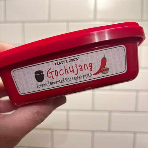Hand holding a red container of Trader Joe’s Gochujang, a Korean fermented red pepper paste, in front of a white subway tile kitchen wall.
