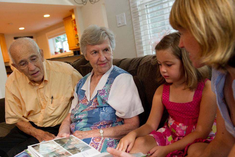 A senior couple sits on a couch with their family, looking through a photo album with a young girl and woman, sharing stories and memories across generations.