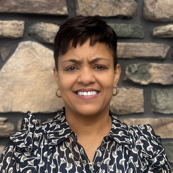 Tracey Coad, Resident Services Director at Vitalia Highland Heights, smiling in front of a stone wall, wearing a patterned black blouse and gold hoop earrings.