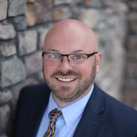 Mark Stapleton, Regional Executive Director at Arrow Senior Living, smiling in a suit and patterned tie while standing beside a textured stone wall.