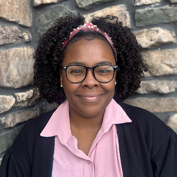 Candice Motley, Wellness Director at Vitalia Highland Heights, smiling in a professional headshot, wearing glasses, a pink blouse, and a black cardigan, with a stone wall background.