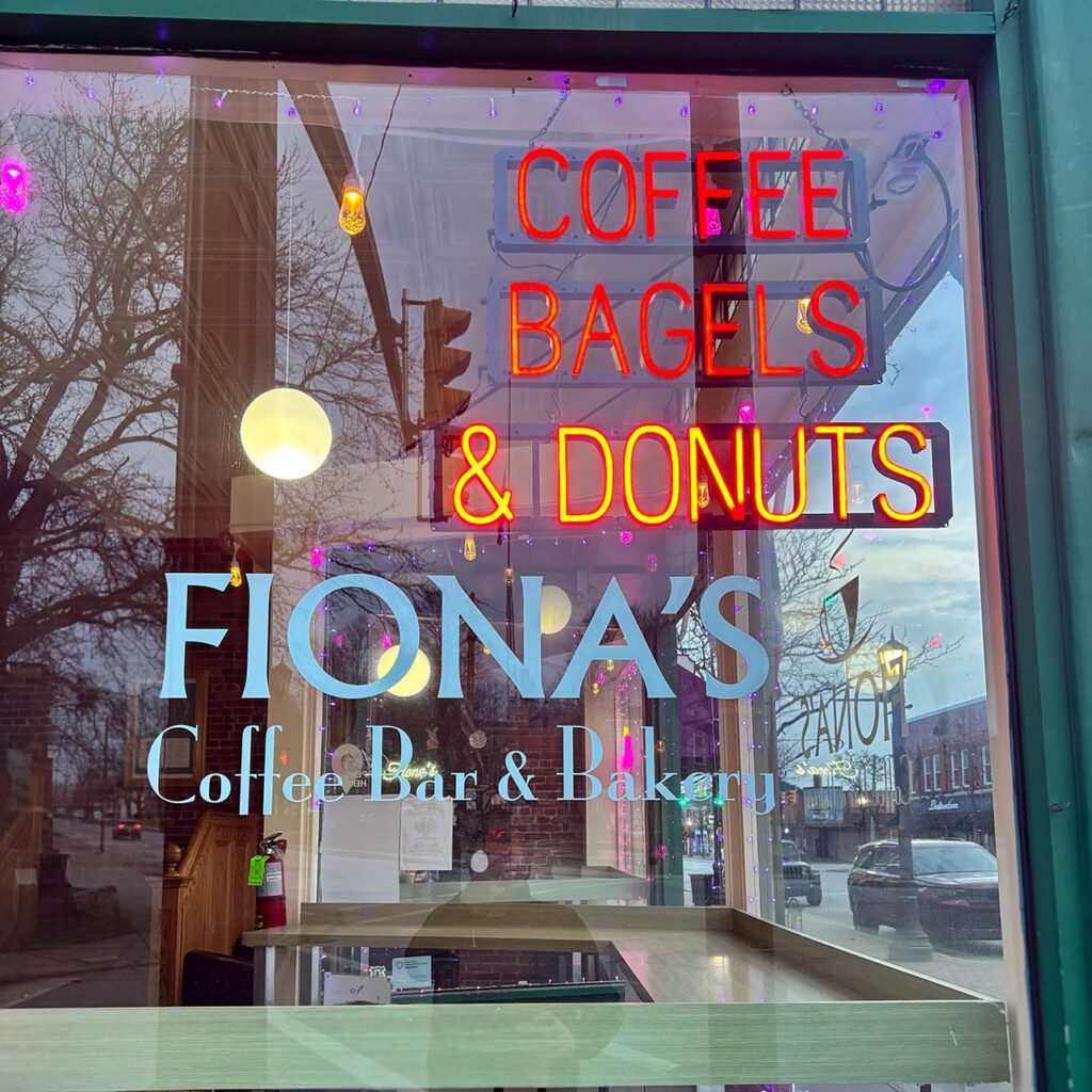 Close-up of the window sign for Fiona's Coffee Bar & Bakery, featuring "Coffee, Bagels & Donuts" lit up in neon.