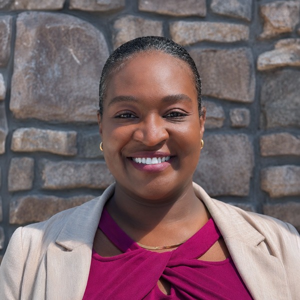 Chanelle Velazquez, Senior Living Director at Vitalia Highland Heights, smiling in a professional headshot with hair pulled back, gold earrings, a magenta blouse, a light blazer, and a stacked stone wall background.