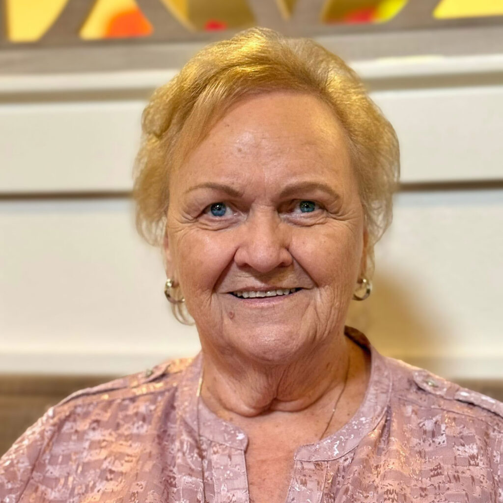 A senior woman wearing a peach blouse smiles welcomingly at Vitalia Highland Heights.