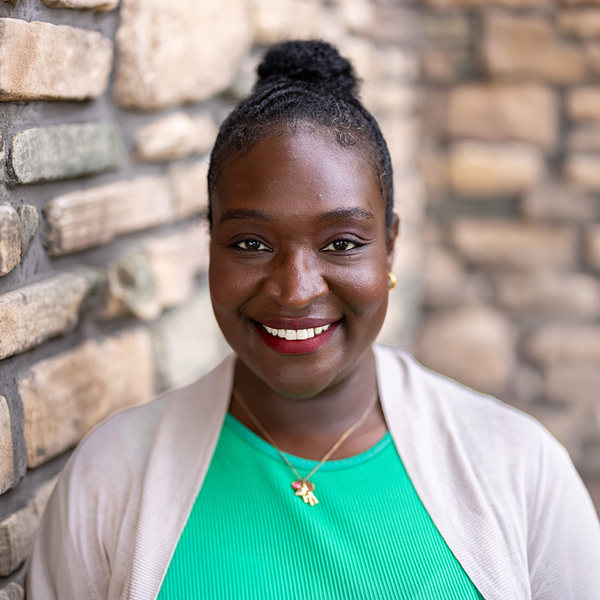 Chanelle Velazquez, Senior Living Director at Vitalia Highland Heights, smiling in a professional headshot with hair in a bun, a green top, a light cardigan, and a softly blurred stone wall background.