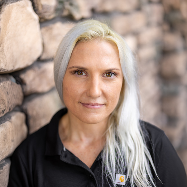 Stephanie Cook, Plant Operations Director at Vitalia Highland Heights, looking at the camera in a professional headshot with long platinum blonde hair, a black polo shirt, and a softly blurred stone wall background.