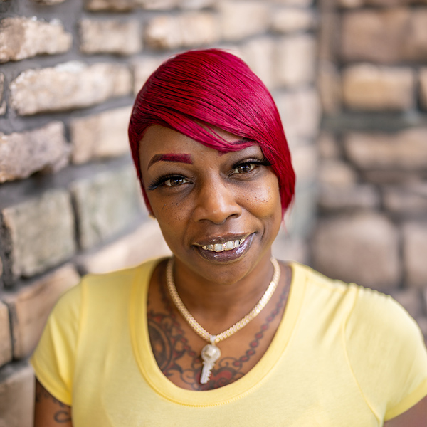 Tomesha Fairley, Memory Care Director at Vitalia Highland Heights, looking at the camera in a professional headshot with bright red short hair, a yellow top, gold jewelry, and a softly blurred stone wall background.
